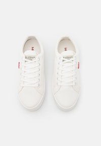 Baskets en toile blanches avec un design à tige basse. Les caractéristiques incluent des lacets ronds, des coutures sur les côtés et une étiquette rouge sur le côté extérieur.