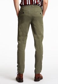 Olijfgroene broeken met een taps toelopend ontwerp, voorzien van twee achterzakken met knopen en een glad, lichtgewicht stof. G gedragen met bruine laarzen.