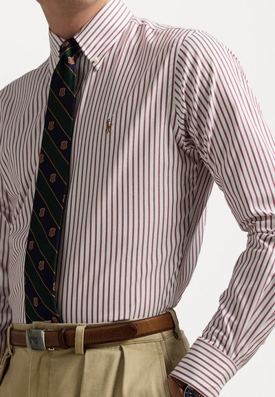 Chemise blanche à rayures rouges étroites, col boutonné. Cravate vert foncé avec des motifs dorés, ceinture marron et pantalon de couleur claire.