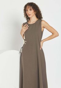 Mujer de cabello rizado con vestido maxi marrón sin mangas con detalle de lazo lateral, llevando pendientes dorados llamativos y anillo floral de gran tamaño.