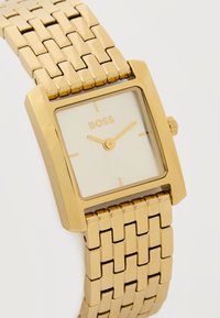 Χρυσό τετράγωνο ρολόι χειρός με συνδεδεμένο λουράκι, μινιμαλιστικό καντράν χωρίς αριθμούς και λογότυπο "BOSS" κάτω από τις 12 η ώρα.