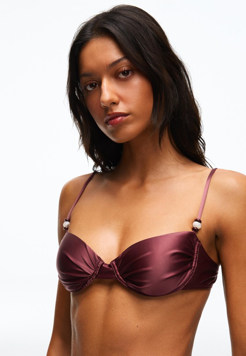 Jeune femme aux longs cheveux foncés portant un soutien-gorge en satin bordeaux avec des bretelles fines et des détails en perles, sur un fond blanc uni.