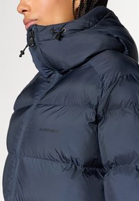 Jachetă puffer de culoare bleumarin, cu guler înalt. Dispune de un fermoar frontal, un șnur ajustabil și un detaliu cu logo pe piept. Textură netedă.