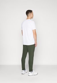 Biela tričko s krátkymi rukávmi, olivovo zelené slim-fit nohavice, biele tenisky s modrými akcentmi. Model je otočený chrbtom k jednému pozadiu.