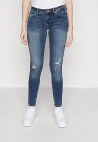 Jeans ajustados de mezclilla azul con detalles desgastados y decoloración. Diseño de cinco bolsillos, cierre de botones y trabillas para cinturón. Combinados con zapatillas blancas.