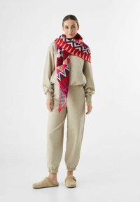 Frau steht in beiger Freizeitkleidung, beigen Slippers und einem großen Schal mit geometrischem Muster in Rot, Pink und Lila, der über die Schultern gelegt ist.