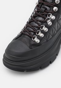 Bota de senderismo de cuero negro con lados acolchados, cordones negros con puntos rojos, ojales plateados, suela de goma gruesa y puntera reforzada.