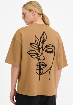 Persona bionda con occhiali da sole e una maglietta beige oversize con un disegno in stile line art nero di un volto e foglie sulla schiena.