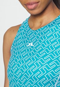 Turkusowa sportowa koszulka na ramiączkach z geometrycznym białym wzorem, okrągłym dekoltem i logo na piersi. Miękki, elastyczny materiał.