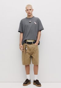 Grå oversized t-shirt med et lille grafisk motiv, beige shorts, farverig mønstret bælte og brune sko. Afsluttet med hvide sokker.