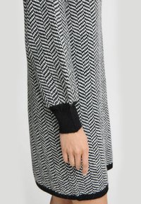 Strickkleid im schwarz-weißen Fischgrätenmuster. Hat lange Ärmel und schwarze gerippte Manschetten mit einem lässigen Schnitt.