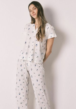 Vrouw met lang haar die een wit pyjama-set draagt, bedrukt met kleine cartoonberen en tekst, staand tegen een effen lichte achtergrond.