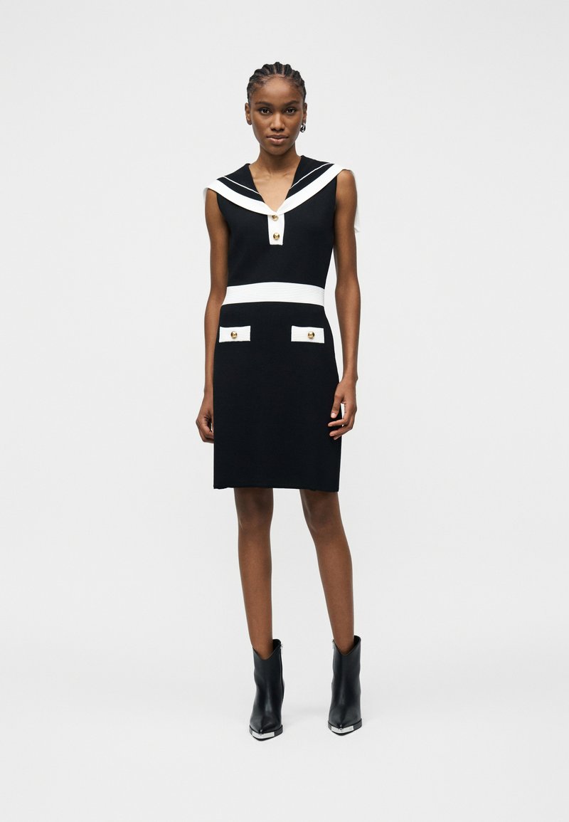 Γυναίκα όρθια, φορώντας ένα αμάνικο μαύρο φόρεμα με λευκή επένδυση, χρυσά κουμπιά και μαύρα μποτίνια μέχρι τον αστράγαλο, μπροστά σε απλό ανοιχτό φόντο.