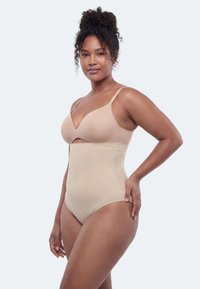 Nő bézs, magas derekú alakformáló fehérneműben és hozzá illő melltartóban, az egyik kezével a csípőjén, világosszürke háttér előtt áll.