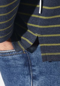 Hand in einer blauen Denim-Jeans-Tasche unter einem navy- und olivgestreiften Hemd mit seitlichen Schlitzen und weißen Kordelzügen.