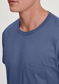 Katoenen T-shirt in een middelblauwe kleur met een ronde hals en een linkerkant borstzakje. De stof heeft een gladde textuur en een subtiel geribbeld ontwerp.