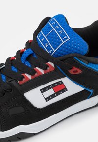 Scarpa sportiva nera con linguetta in rete blu, dettagli rossi e rifiniture bianche. Presenta un logo "TOMMY JEANS" sul lato. Tessuto testurizzato.