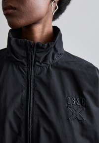 Chaqueta de nylon negra con cuello alto, cremallera frontal y logotipo bordado "032C X" en el pecho. Textura suave, diseño minimalista.