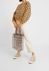 Mujer con sudadera de color beige con estampado, pantalones crema y zapatillas altas beige, sosteniendo un gran bolso de tote transparente con cuadros por las asas.