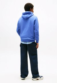 Blauer Kapuzenpullover mit Kordelzug, kombiniert mit einem karierten Hemd und breiten dunkelblauen Hosen. Die Sneaker zeigen eine Mischung aus Weiß und Marineblau.