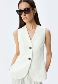 Weißer ärmelloser Blazer mit V-Ausschnitt und zwei schwarzen Knöpfen, mit taillierter Passform und seitlichen Taschen. Schwarze Katzenaugen-Sonnenbrille als Akzent.