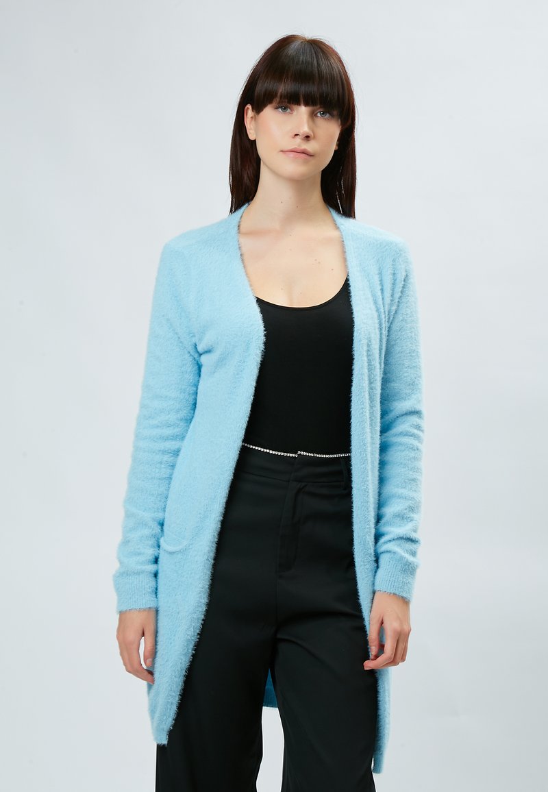 Cardigan bleu clair, duveteux, avec une silhouette longue, une ouverture devant et deux poches avant. Poignets et ourlet côtelés. Porté sur un haut noir.