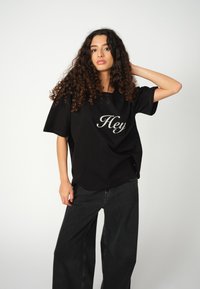 Joven con cabello rizado que lleva una camiseta negra extragrande con la impresión "Hey" y jeans negros holgados, posando con una mano detrás de la cabeza.