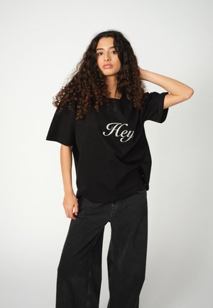 Noor on curly hair wearing a black oversized shirt with "Hey" print and loose black jeans, posing with one hand behind her head.