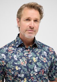 Camicia a maniche corte con fantasia floreale, caratterizzata da uno sfondo blu scuro e da motivi botanici multicolori, con chiusura a bottoni e colletto classico.