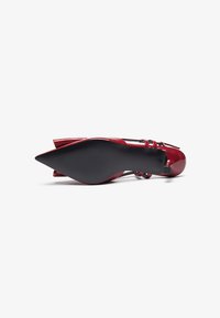 Red patent leather high-heeled shoe with a pointed toe, thin ankle strap, and a textured black sole. Featured bow accent at the heel.