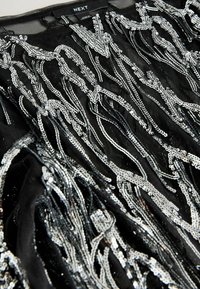 Czarny top z misternymi srebrnymi cekinami w falistych wzorach, wykonany z lekkiego materiału o strukturalnym wykończeniu.