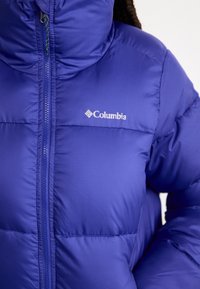 Jachetă puf purpurie, confecționată dintr-un material ușor, cu umplutură matlasată. Are un guler înalt și închidere cu fermoar, cu un logo alb Columbia pe piept.