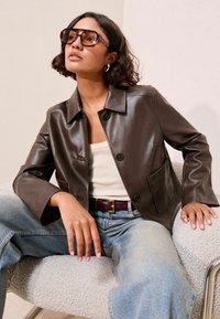 Veste cropped en cuir marron avec un col et une poche avant, associée à un jean en denim bleu clair et un débardeur crème.