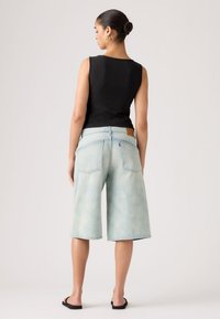 Γυναίκα που στέκεται με την πλάτη γυρισμένη, φορώντας μαύρο αμάνικο τοπ, ανοιχτό μπλε τζιν σορτς μέχρι το γόνατο, μαύρα σανδάλια slide και χρυσούς κρίκους στα αυτιά.