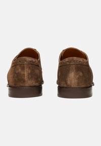 Chaussures en daim marron avec une texture lisse, doublure en cuir et un petit talon marron. Présentent des perforations décoratives le long de l'arrière.