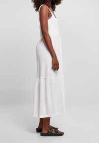Biała maxi sukienka z regulowanymi ramiączkami, z warstwowym dołem i lekką, gładką teksturą. Noszona z ciemnymi sandałami.