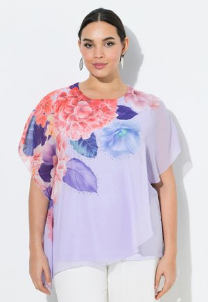 Frau trägt eine hellviolette Bluse mit großen roten, rosa und blauen Blumenmustern, kombiniert mit weißen Hosen vor einfachem Hintergrund.