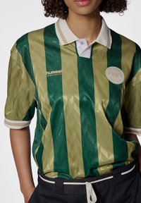 Grøn- og guldstribet polo-shirt med et tekstureret mønster, en hvid krave og "Hummel Sportswear" logo på brystet. Sort bæltedragt synlig.