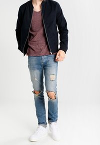 Man wearing a black jacket, maroon t-shirt, ripped blue jeans, and white sneakers, standing against a plain white background.