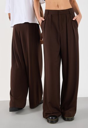 Brown wide-legged trousers with pleats, made of a smooth fabric. Features side pockets and paired with chunky black footwear.