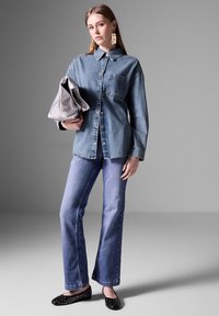 Camicia in denim con colletto e tasca, abbinata a jeans a zampa. Tiene una borsa a tracolla argentata. Ballerine nere con borchie. Sfondo semplice grigio.