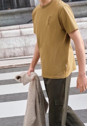 Persoon die een mosterdgele T-shirt met geborduurde "f" draagt en een beige gebreide trui vasthoudt, staand bij de stoeprand van een zebrapad.