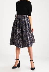 Námořnická sukně s květinovým potiskem a lesklým metalickým vzorem, ke kolenům, s nařaseným pasem, spárovaná s černým topem a podpatky. Hladká textura.
