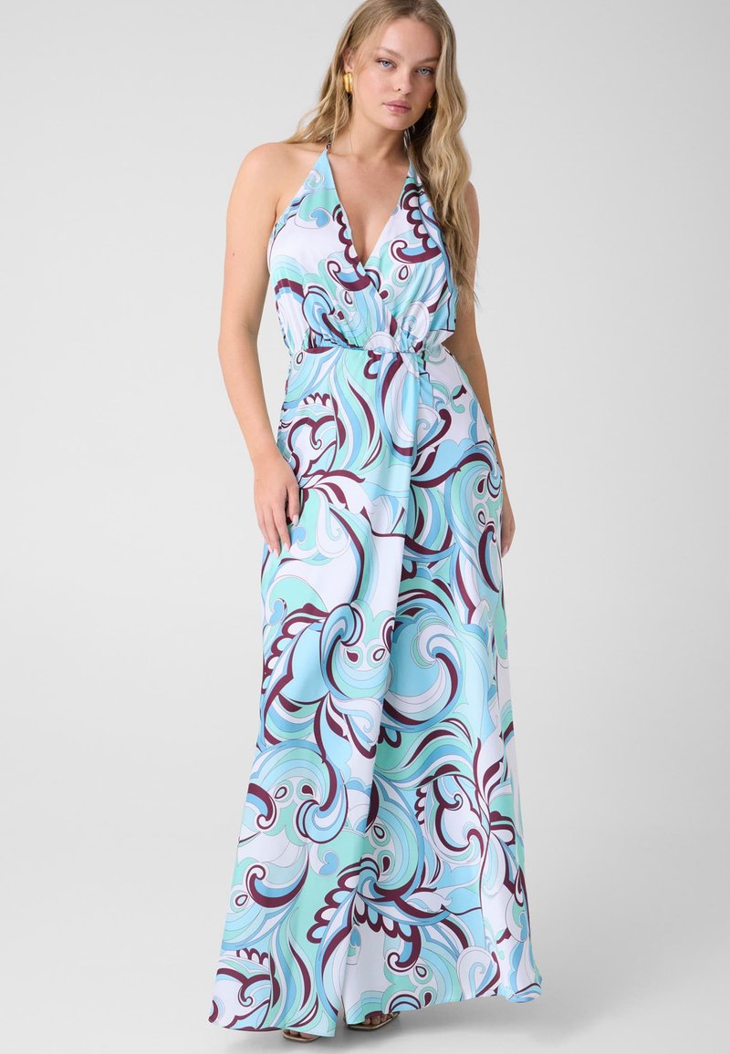 Femme portant une robe maxi sans manches, col halter, avec un motif tourbillonnant bleu, blanc et violet, debout devant un fond uni.