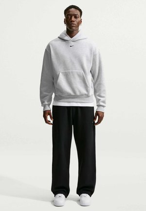 Νεαρός άνδρας φοράει ανοιχτό γκρι φούτερ Nike, μαύρο παντελόνι και λευκά αθλητικά, στέκεται μπροστά σε απλό λευκό φόντο.