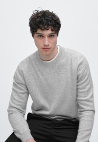 Jasnoszary dzianinowy sweter z okrągłym dekoltem, miękka faktura, ściągacze na rękawach, noszony na białej koszuli. Model siedzi na neutralnym tle.