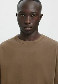 Pruun puuvillane T-särk ümar kaelusega ja lühikeste varrukatega, sileda tekstuuriga ja lõdva istuvusega, mida kannab modell heledal taustal.