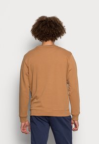 Uomo con capelli ricci che indossa una felpa beige a maniche lunghe e pantaloni blu scuro, in piedi di fronte a una parete grigia chiara e uniforme.