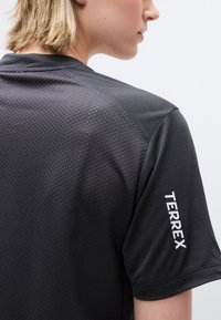 Κοντινό πλάνο ενός ατόμου που φοράει μαύρο κοντομάνικο πουκάμισο με υφή υφάσματος και το λογότυπο "TERREX" τυπωμένο στο μανίκι.