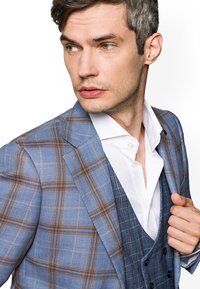 Jasnoniebieska kurtka w kratę z brązowymi akcentami, noszona na białej koszuli i niebieskiej kamizelce. Gładka faktura, wyraźne klapy, dopasowany fason.